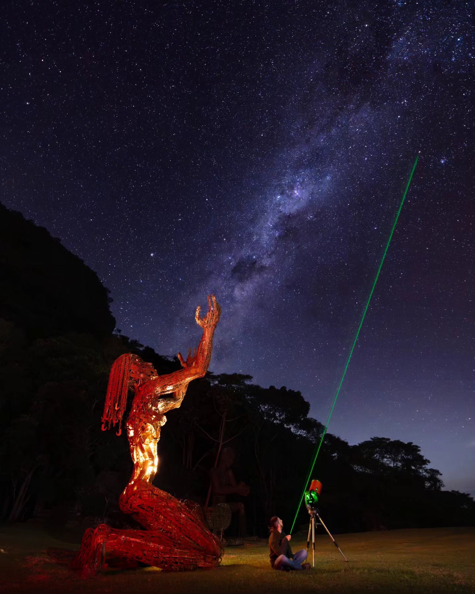 Escultura iluminada sob a Via Láctea, com telescópio apontando um laser ao céu