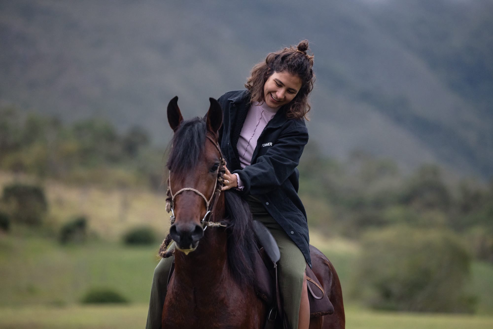 Mulher sorrindo a cavalo, afagando o pescoço do animal, com a serra ao fundo