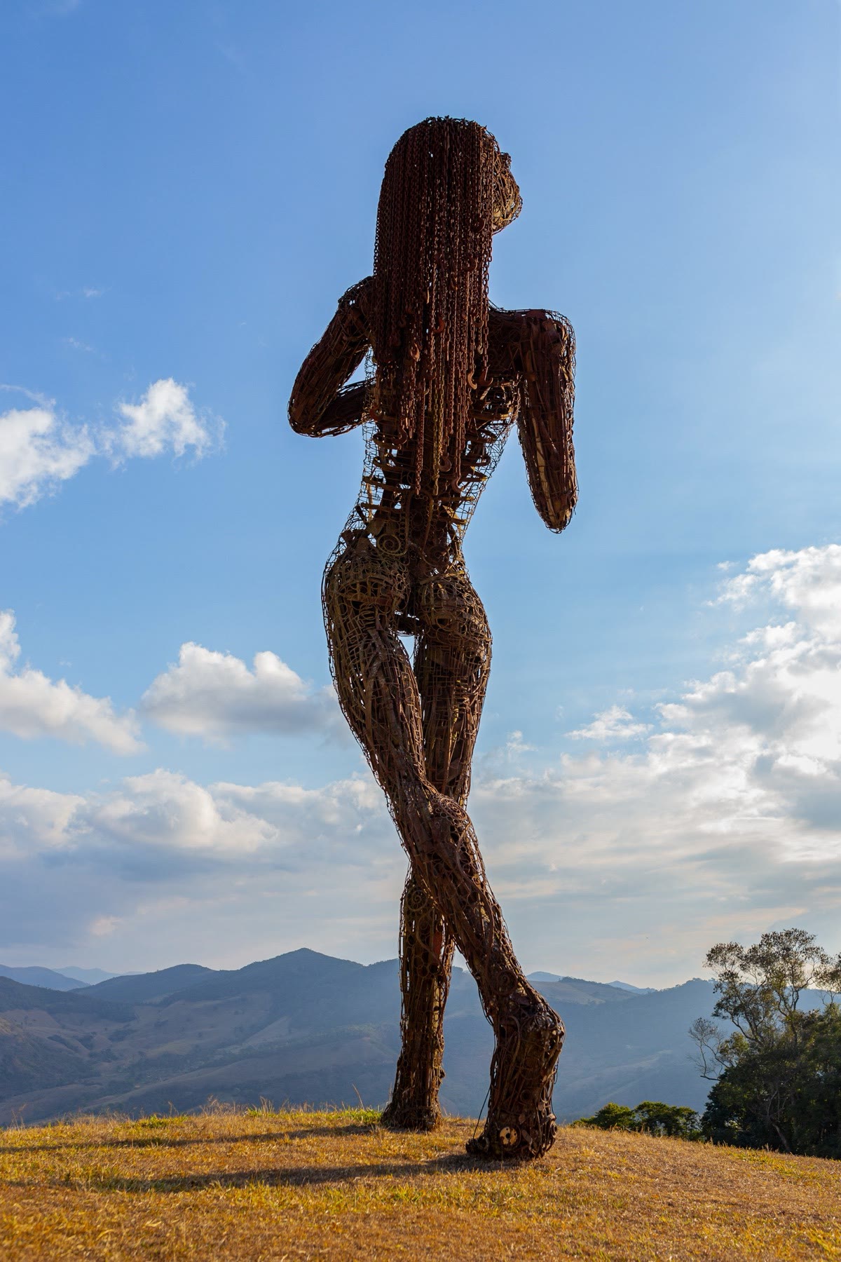 Escultura de ferro em forma de figura humana no alto do mirante, com a serra ao fundo