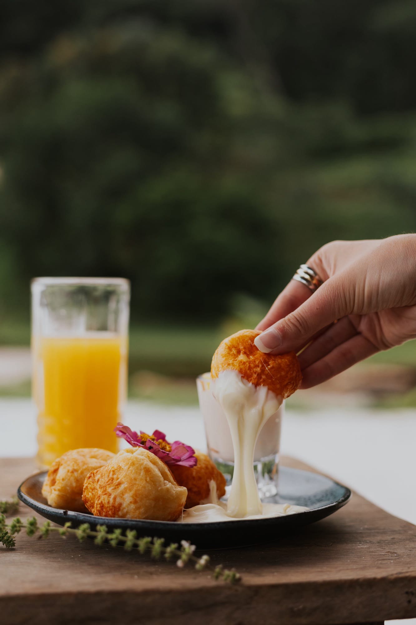 Mão mergulhando um pão de queijo em copinho de molho cremoso, com suco de laranja e flor comestível no prato